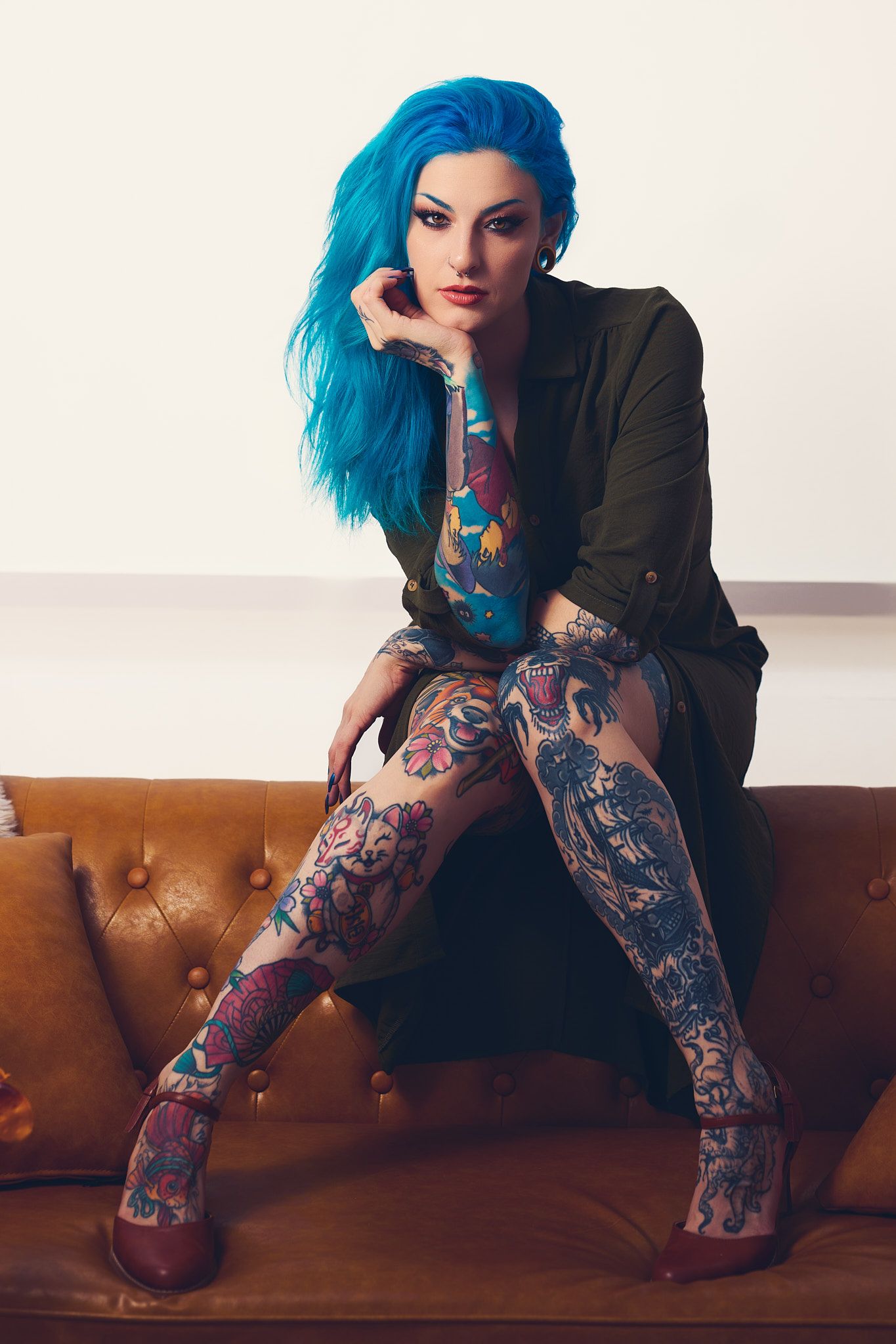 Alternative model with bright blue hair poses on brown leather couch wearing black blazer and patterned tights.