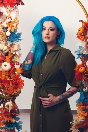 Fashion model in olive green dress poses with vibrant autumn leaves and bright blue hair against white background.