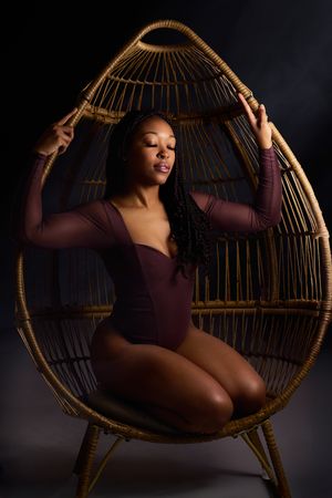 Artistic boudoir photo series showing figure posing gracefully in wicker egg chair against dark background.