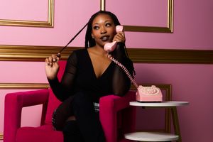 A stylish woman in a black dress poses on a pink chair while talking on a retro telephone.