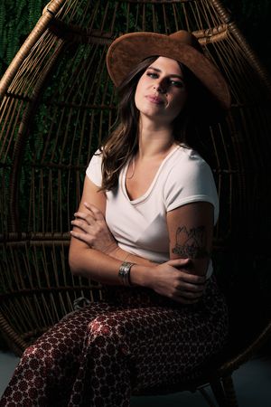 A person in a brown hat and white t-shirt sits in a wicker chair wearing patterned pants and displaying arm tattoos.