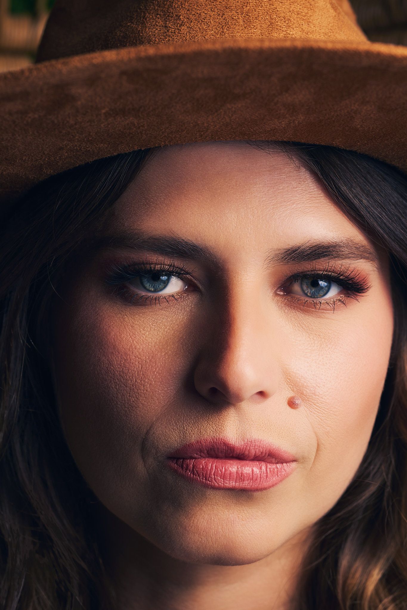 Close-up portrait of a woman wearing a brown hat with dramatic lighting highlighting her striking blue eyes and red lips.