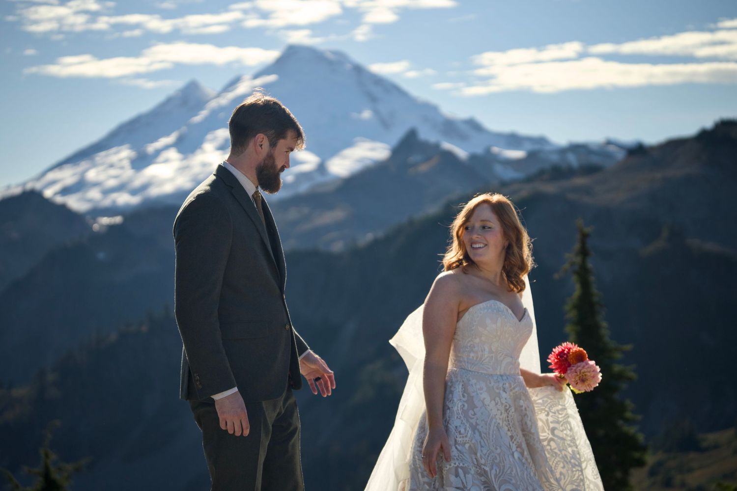 A smiling bride and groom walk together in front of a majestic, snow-capped mountain peak under a clear blue sky.