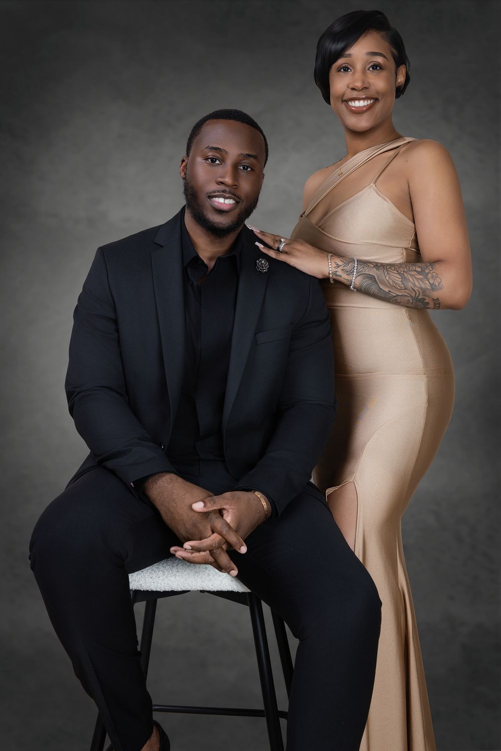 Professional studio portrait featuring a couple in formal attire with black suit and beige dress against gray backdrop.