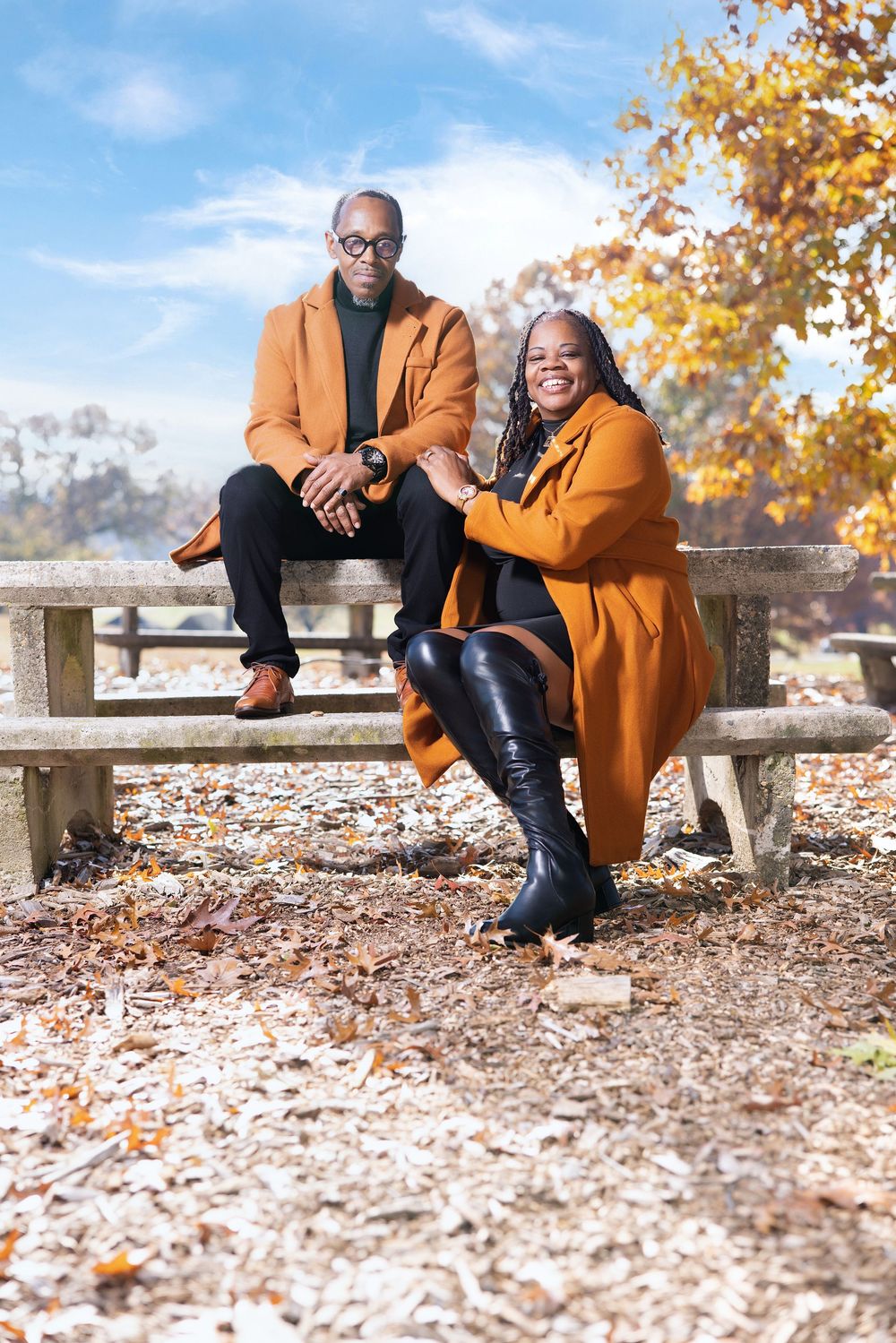 A couple sits together on a wooden bench surrounded by fallen autumn leaves on a bright sunny day.