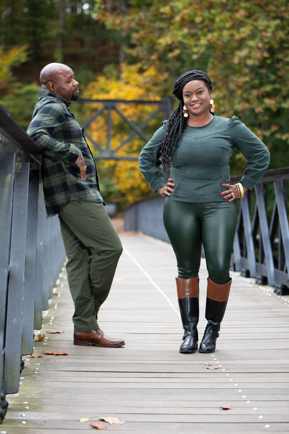 Couple in matching green outfits poses playfully on wooden bridge during autumn photo session.