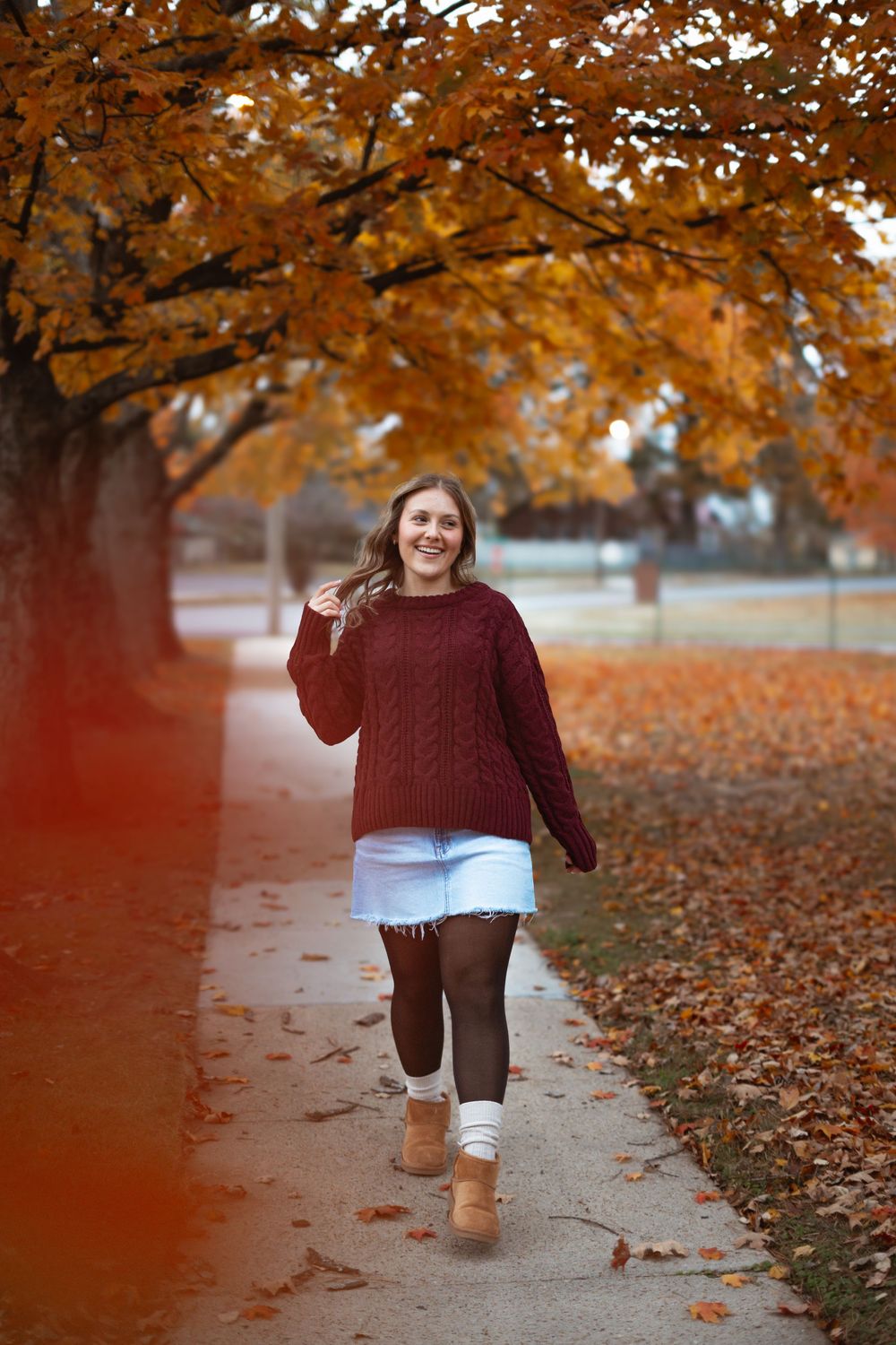 Someone in a maroon sweater walks through fallen autumn leaves under orange-colored trees.