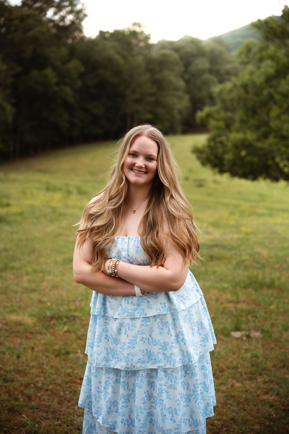 A light blue floral dress flows in a sunny meadow surrounded by green trees and grass.