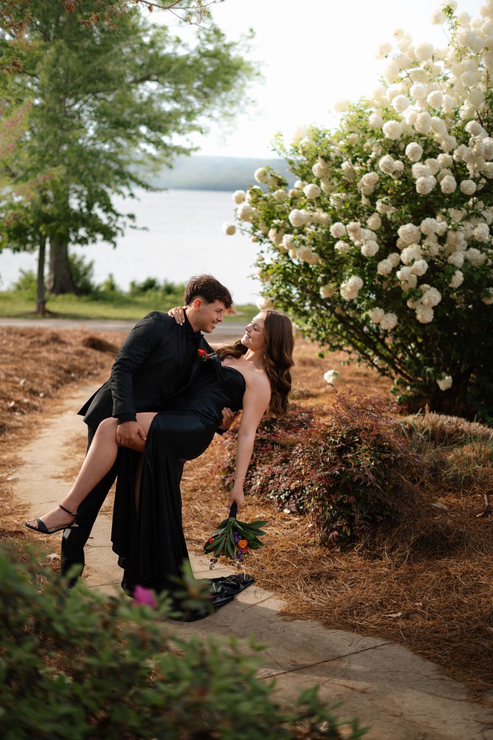 A couple poses dramatically among blooming white roses near a waterfront.