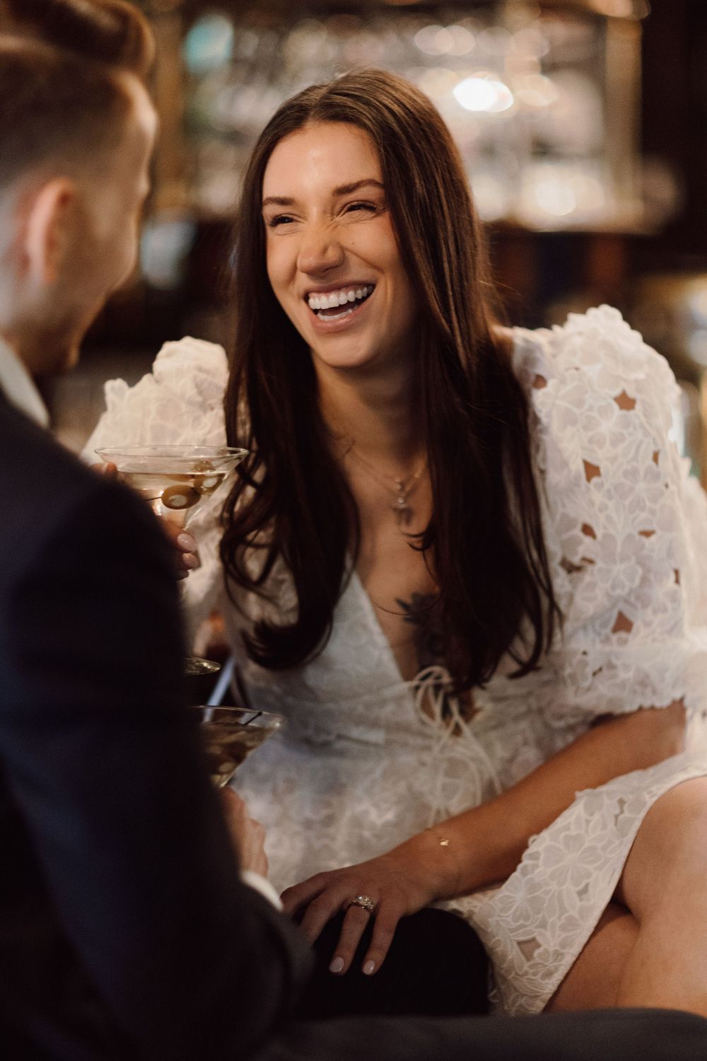 Woman in white lace dress laughing at a bar counter.