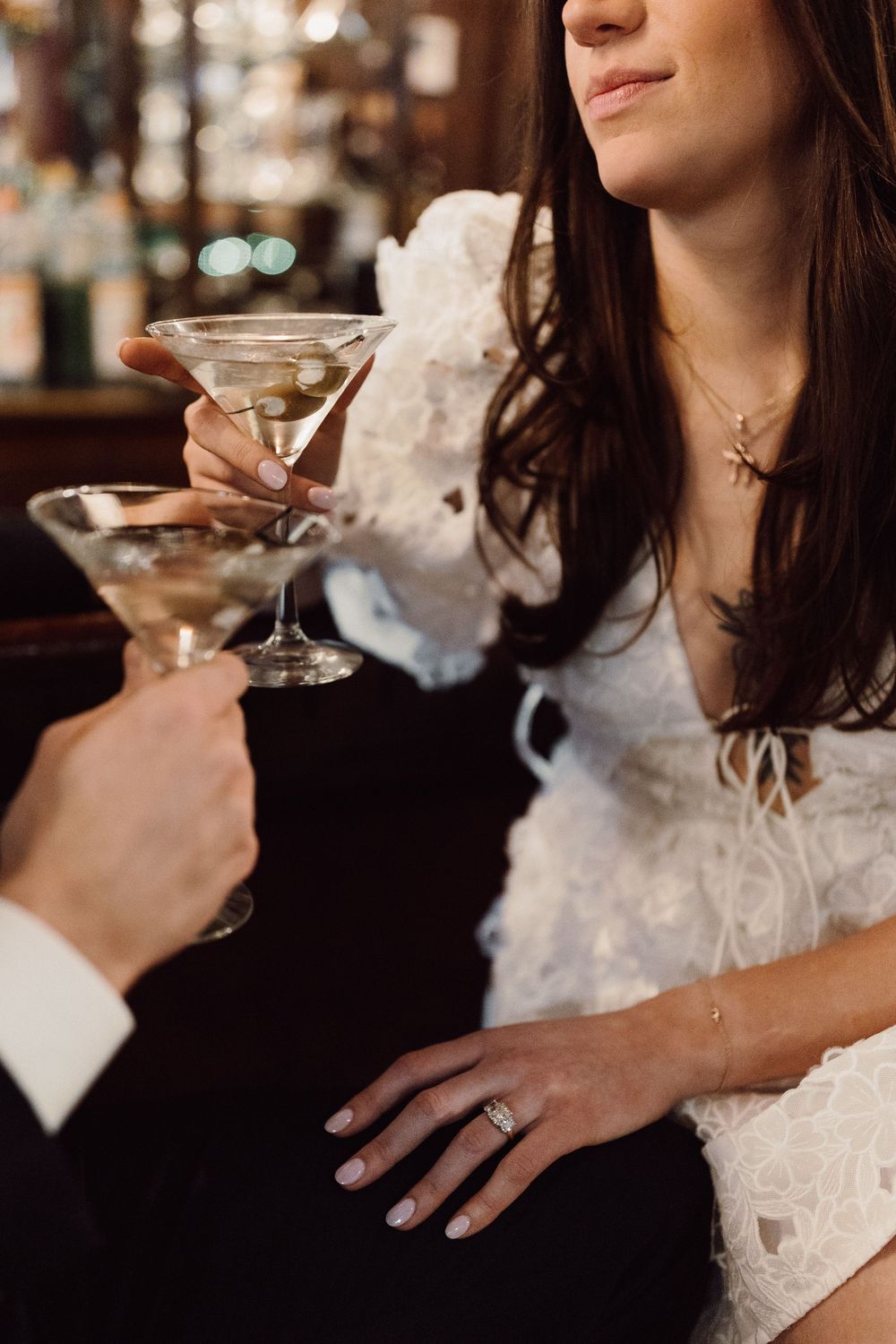 Two martini glasses clinking in celebration at a bar with white lace dress details visible.