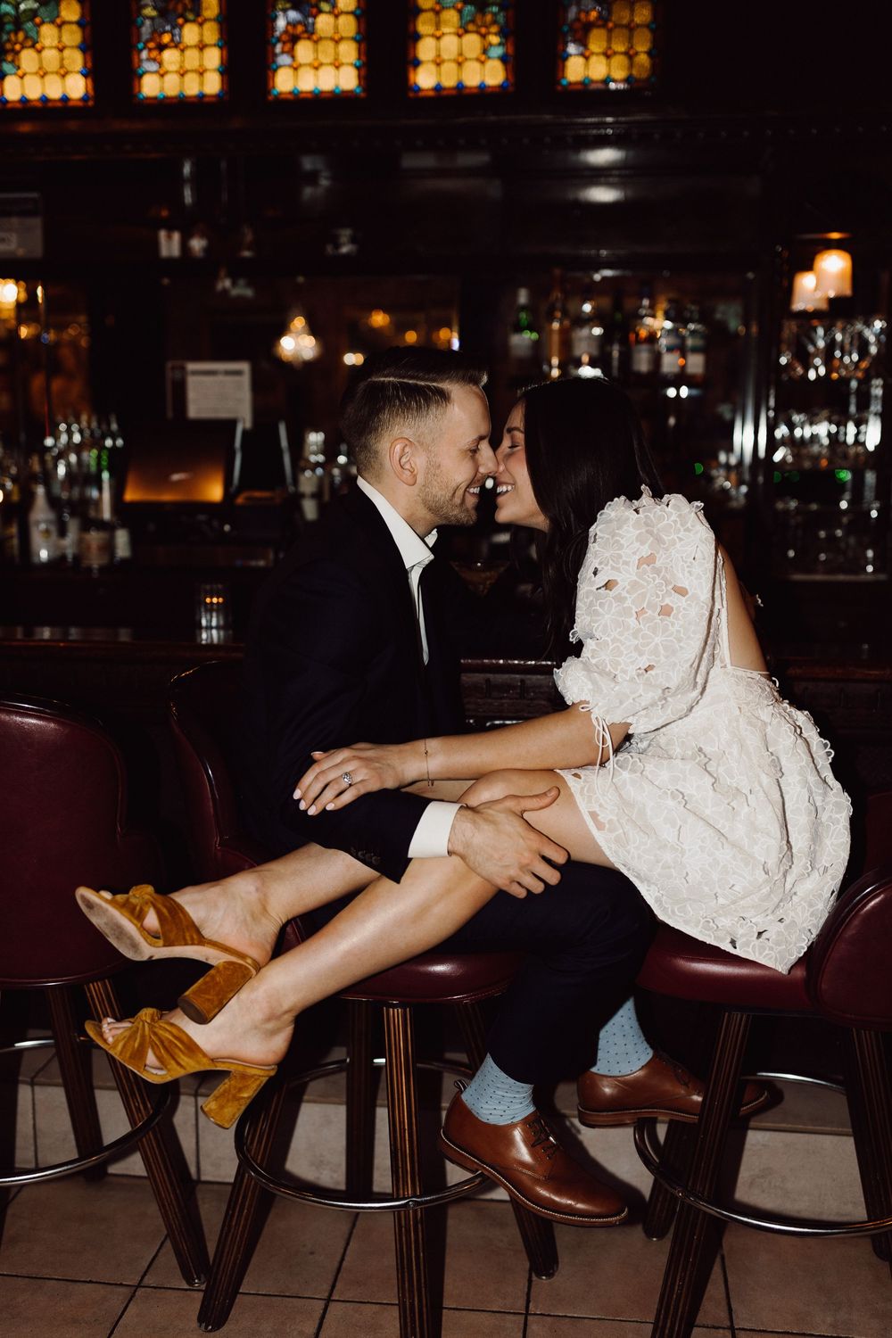Couple in formal attire sharing intimate moment at vintage bar with stained glass windows.