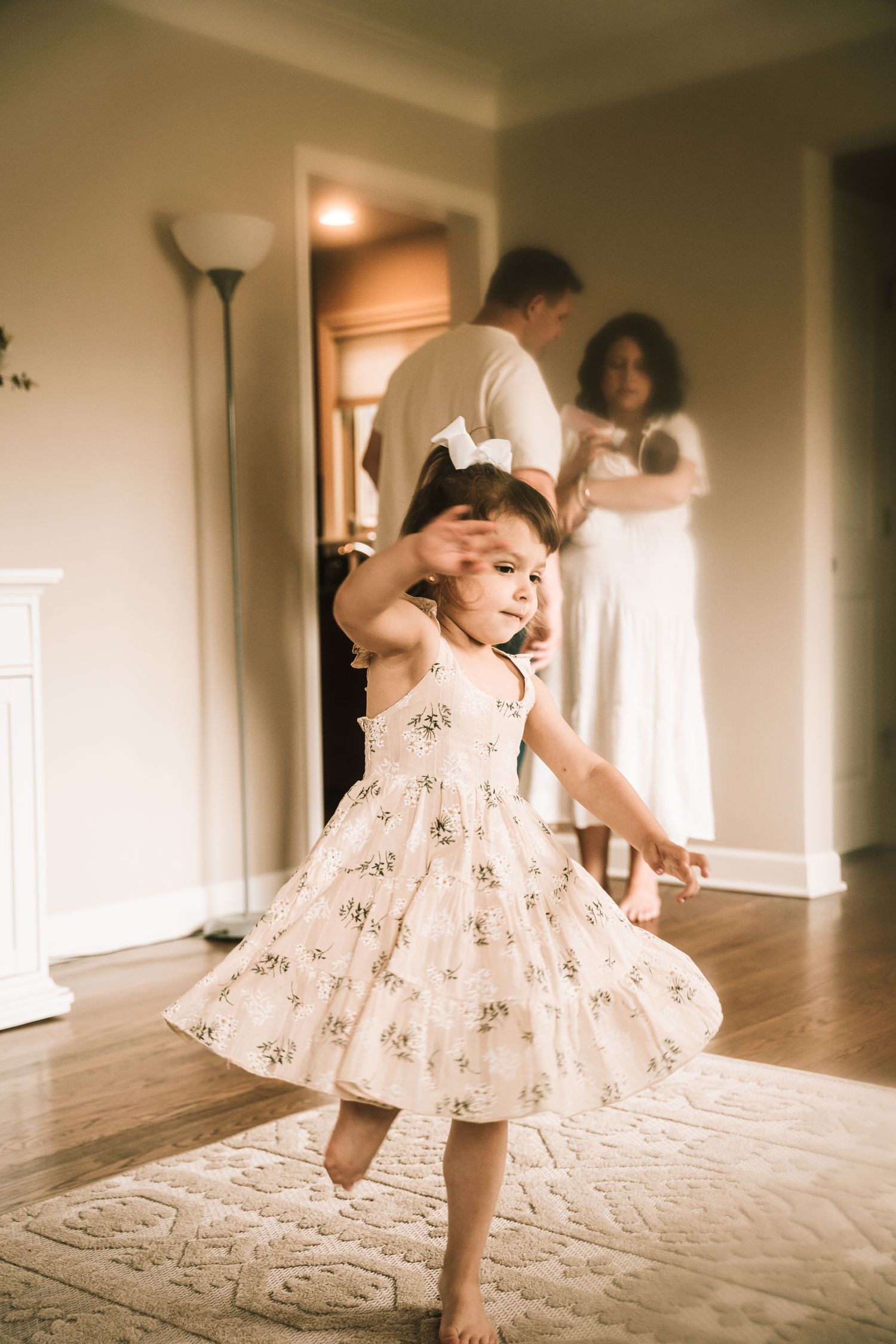 A young child in a white floral dress twirls and dances in a warmly lit living room while recording a family film.