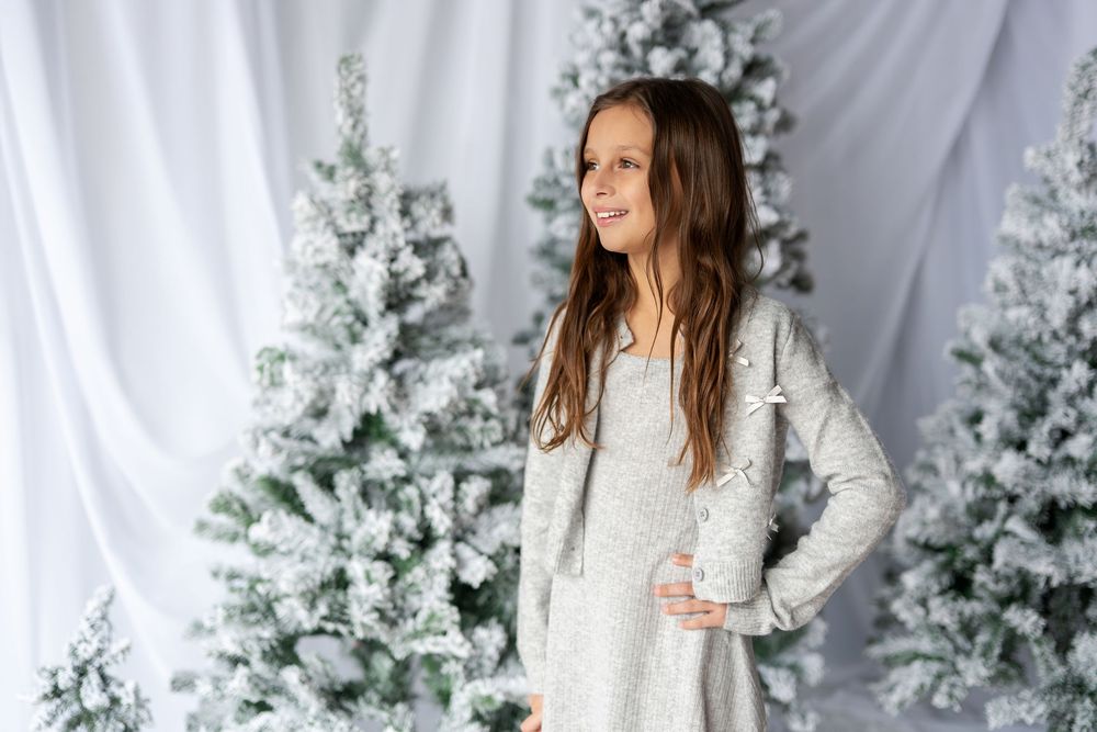 Cheerful young girl in a gray long-sleeve dress posing beside flocked white Christmas trees in a bright studio.