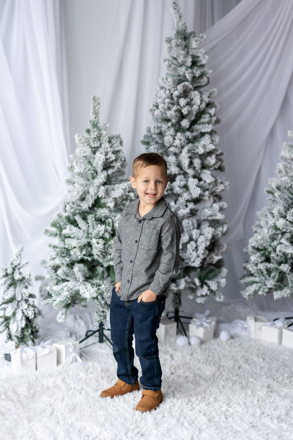 Smiling young boy in a gray shirt and jeans standing in front of snow-flocked Christmas trees with wrapped gifts.