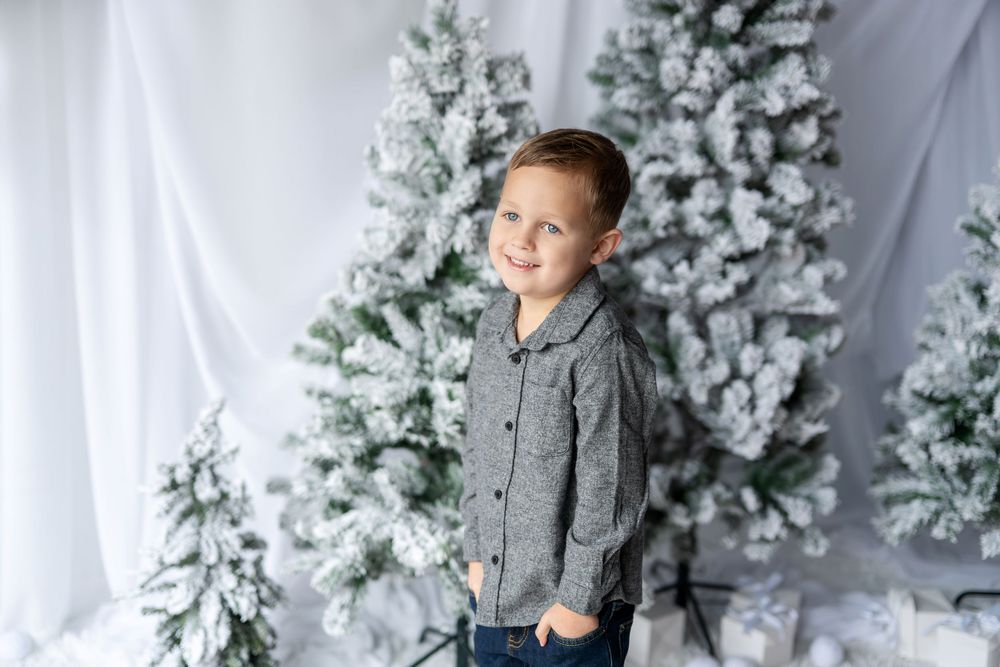Young boy in a gray jacket and jeans posing beside tall flocked Christmas trees in a white holiday studio.
