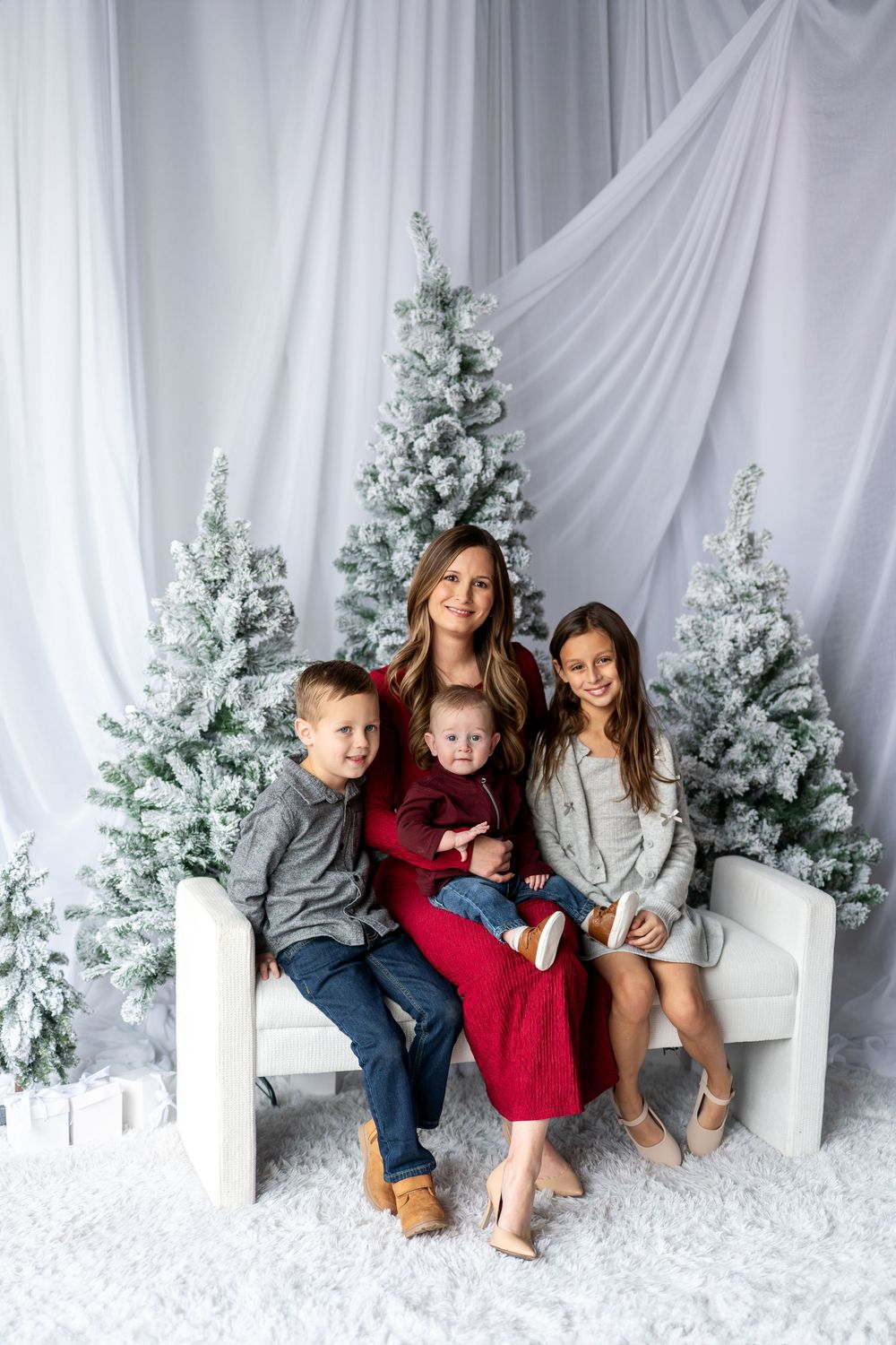 A smiling mother in a red dress sits with her three children on a white bench beside frosted holiday trees.