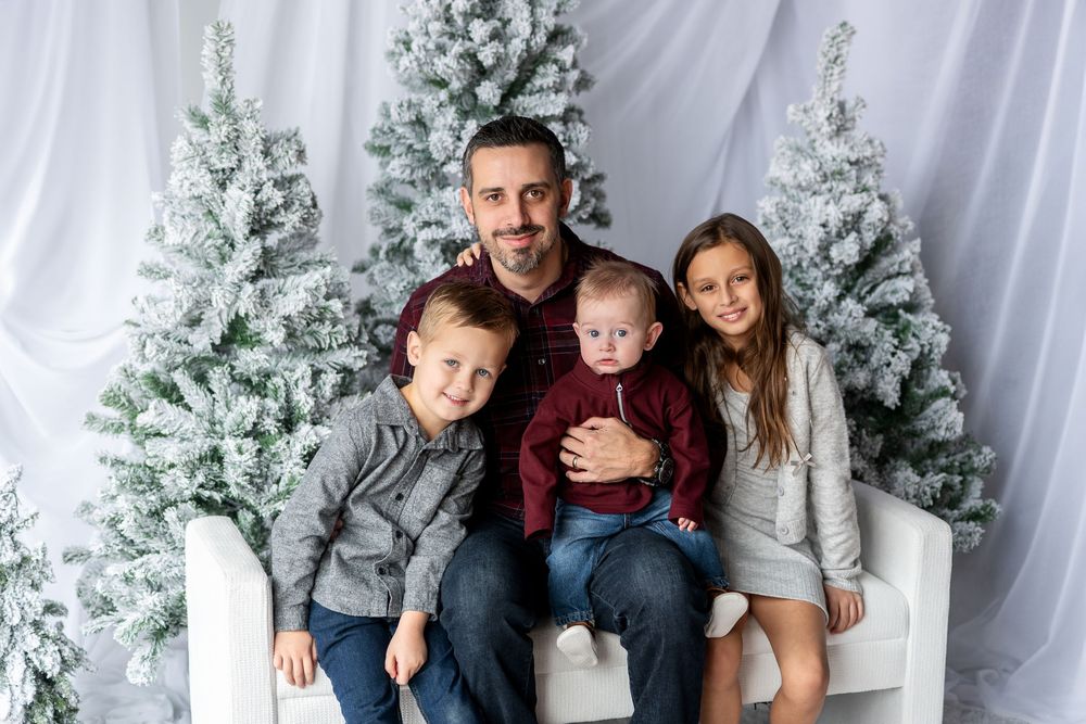 A father in burgundy sits with his three children on a white bench surrounded by snow-flocked Christmas trees.