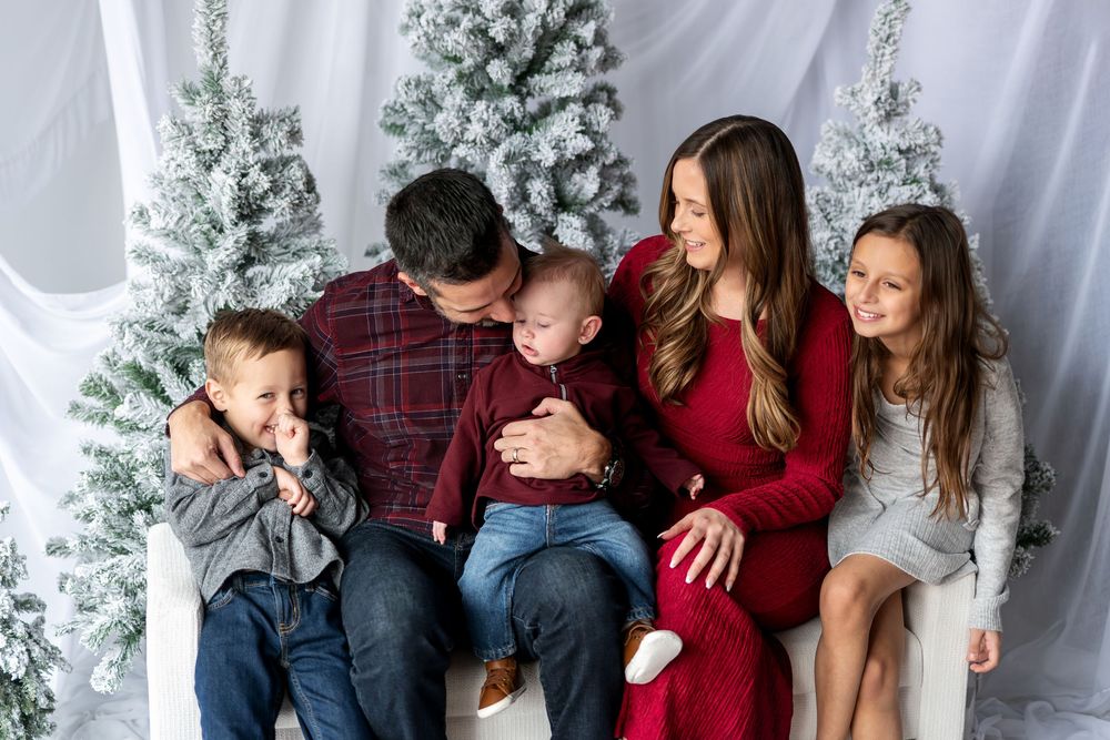 A loving family of five shares a tender moment on a white bench beside frosted Christmas trees.
