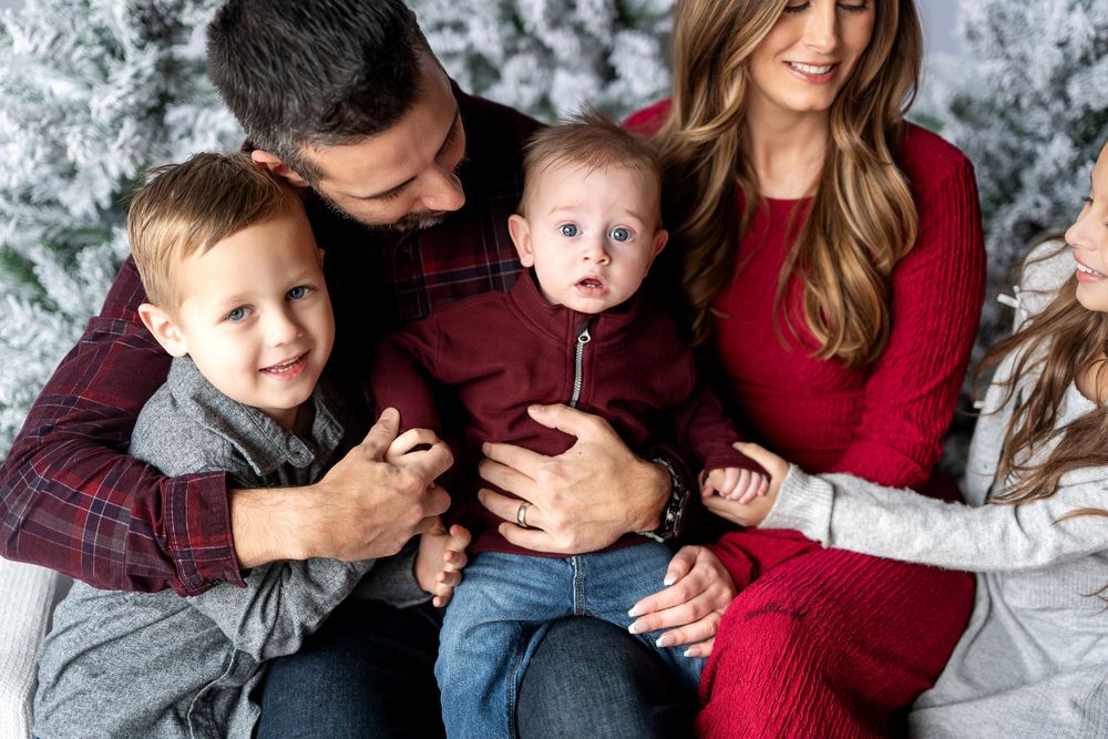 A close-up of a family of five gathered together with snow-flocked Christmas trees in the background.