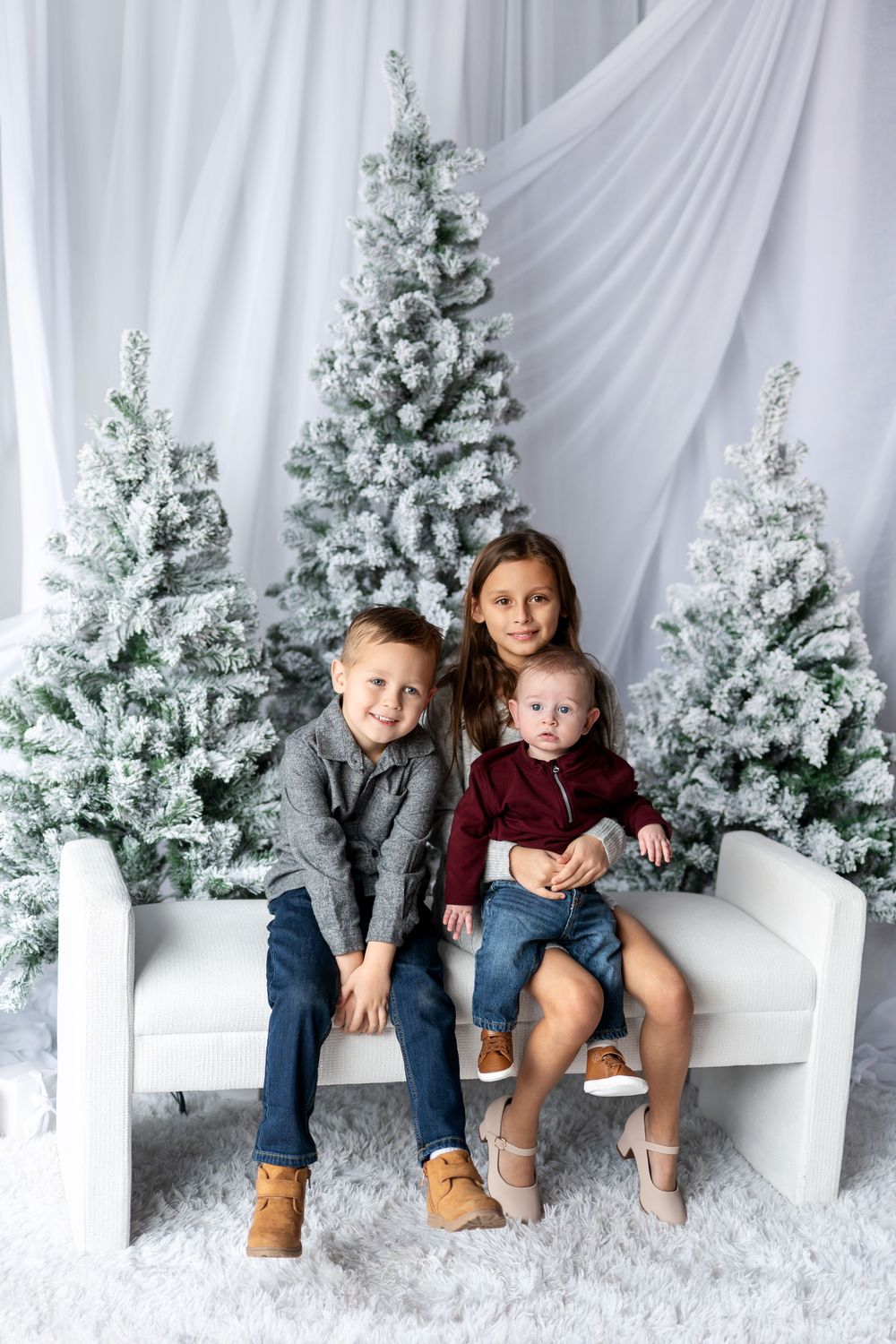 Three siblings sit together on a white bench surrounded by snow-flocked Christmas trees in a holiday photo.