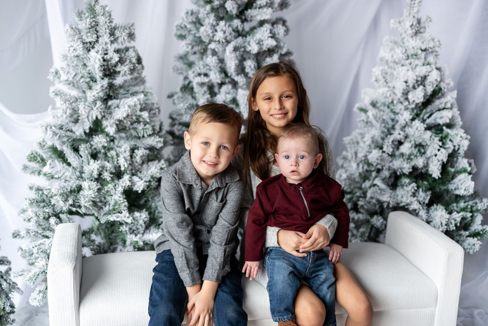 Three siblings pose together on a white bench surrounded by snow-covered Christmas trees in a studio.