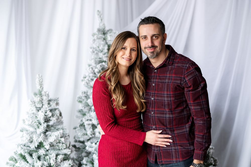 A smiling couple embraces in front of snow-flocked Christmas trees in a festive holiday portrait.