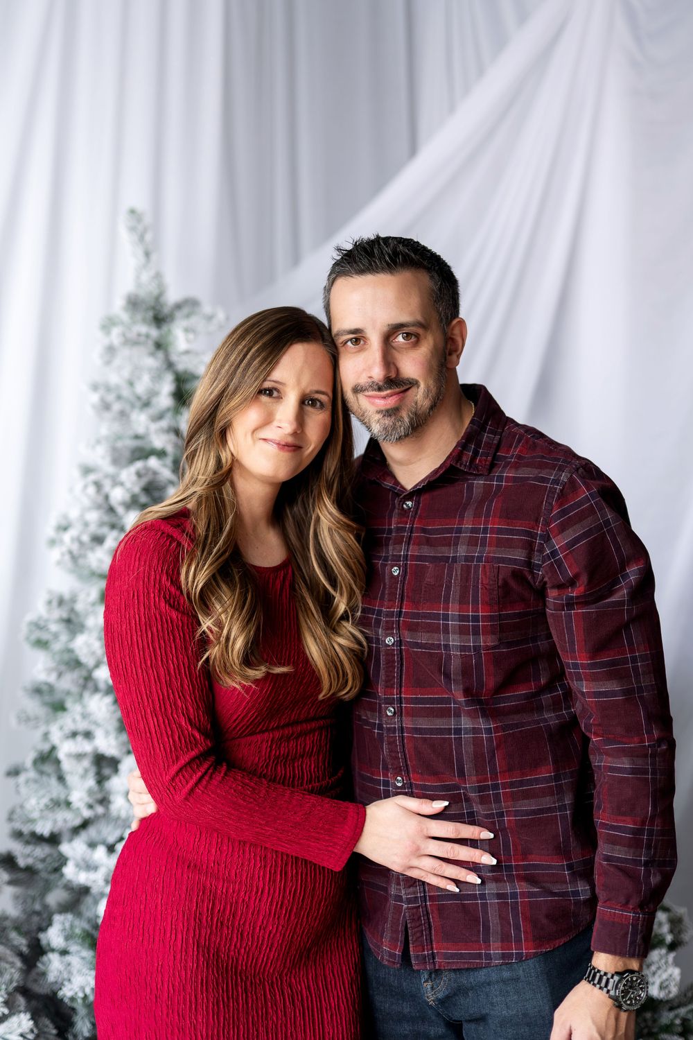 Smiling couple posing together for a Christmas portrait, woman in red knit dress and man in burgundy plaid shirt.