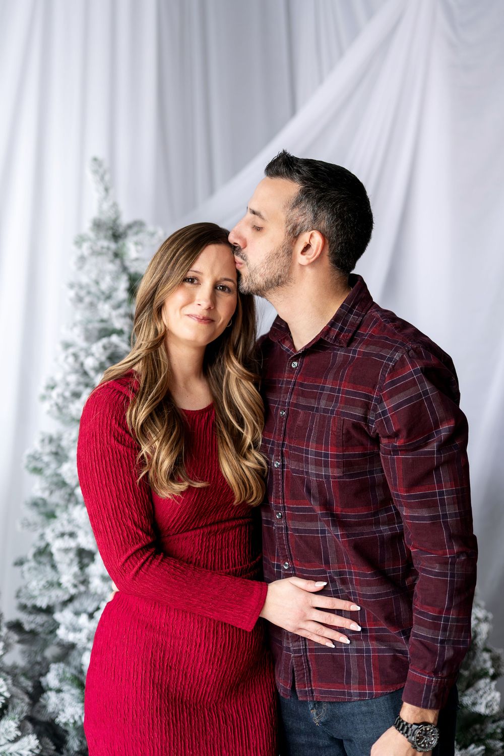 Man tenderly kissing woman on the forehead during a romantic Christmas photo session with snowy trees.