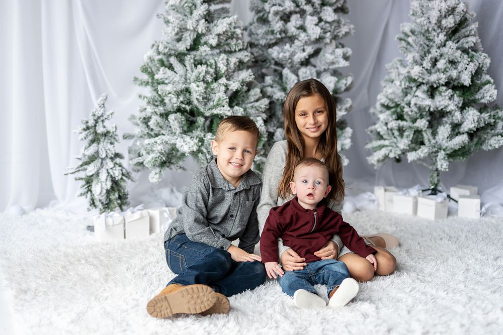 Three siblings sit together on white fluffy carpet surrounded by snow-flocked Christmas trees and gift boxes.