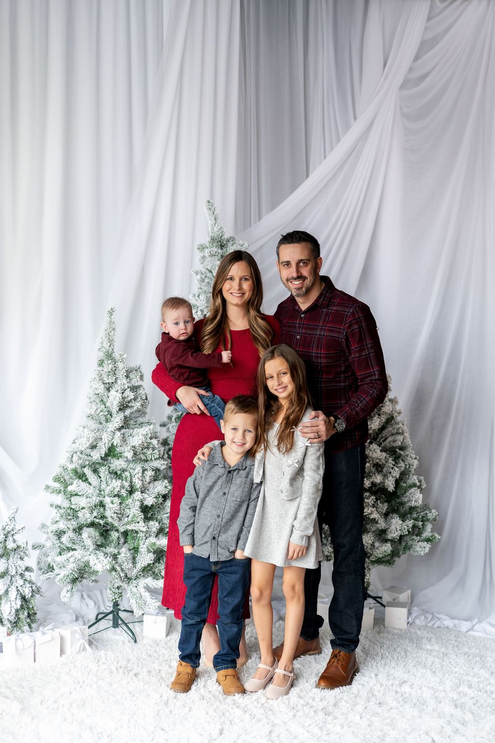 Happy family of five smiling together in front of snow-flocked Christmas trees in a bright white studio setting.