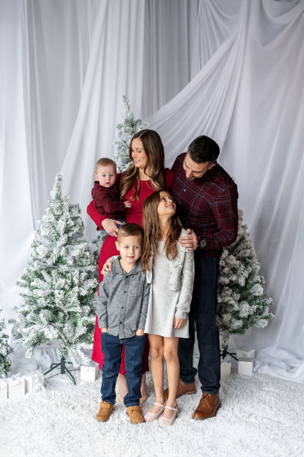 Family of five sharing a tender moment by white flocked Christmas trees, dressed in coordinating red and gray holiday outfits.