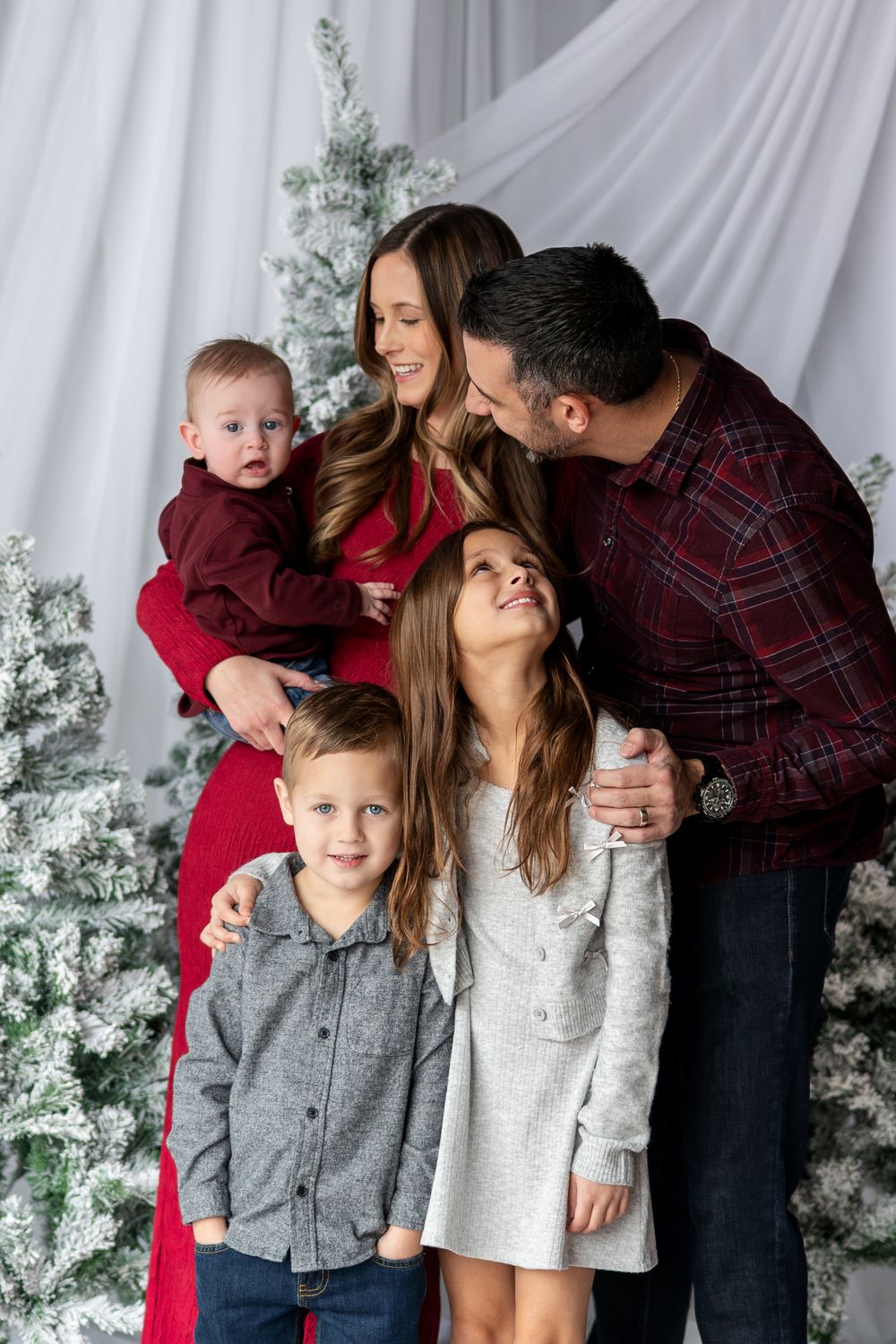 Family of five posing playfully by flocked Christmas trees, parents in red and plaid with three children in festive attire.