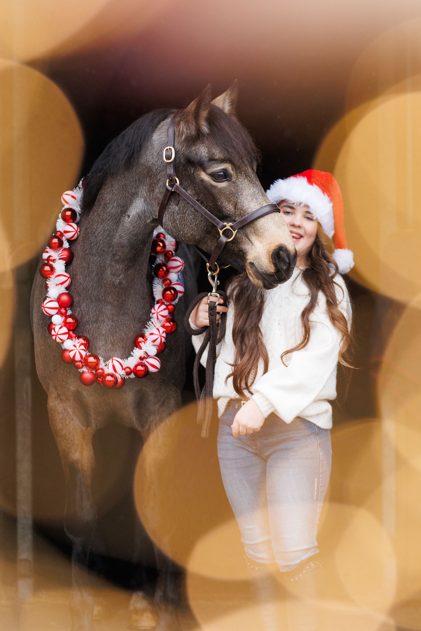 A brown horse and rider in a white sweater pose together against autumn foliage in soft natural lighting.