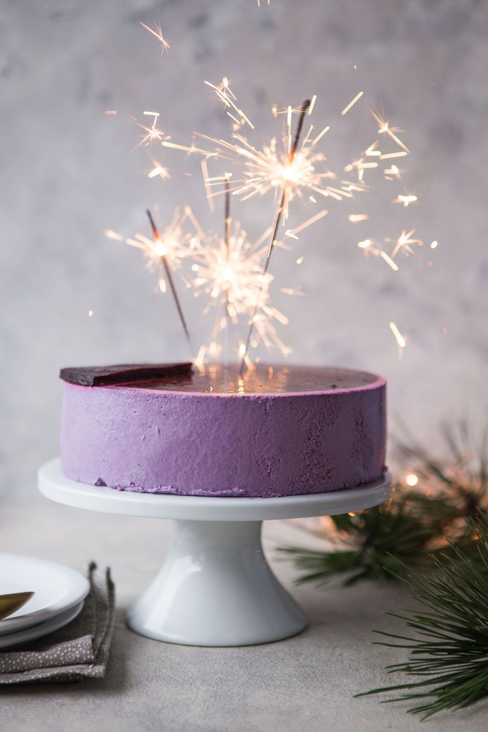A purple cheesecake on a white cake stand decorated with sparklers creating a festive display.