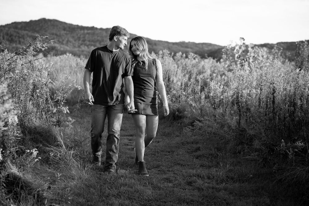 A couple walks together on a dirt trail through wild grasses with hills in the background, captured in black and white.
