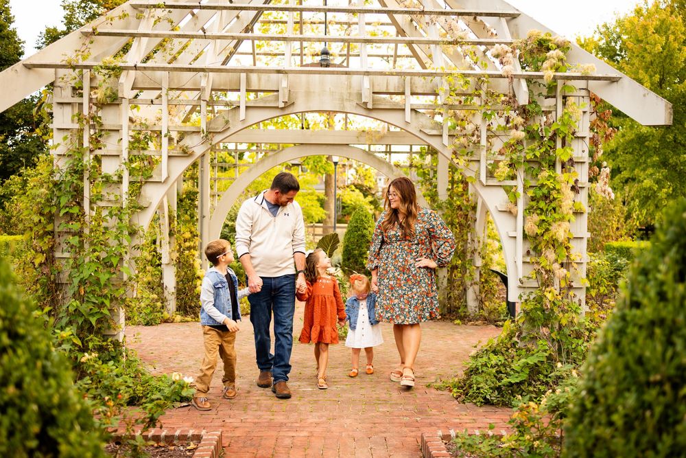 A family walks through a rustic garden arbor pathway surrounded by lush greenery and brick pavers.