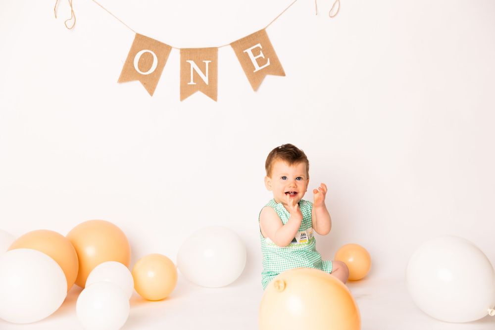 Baby in mint green outfit sits among white and peach colored balloons for first birthday photo session.