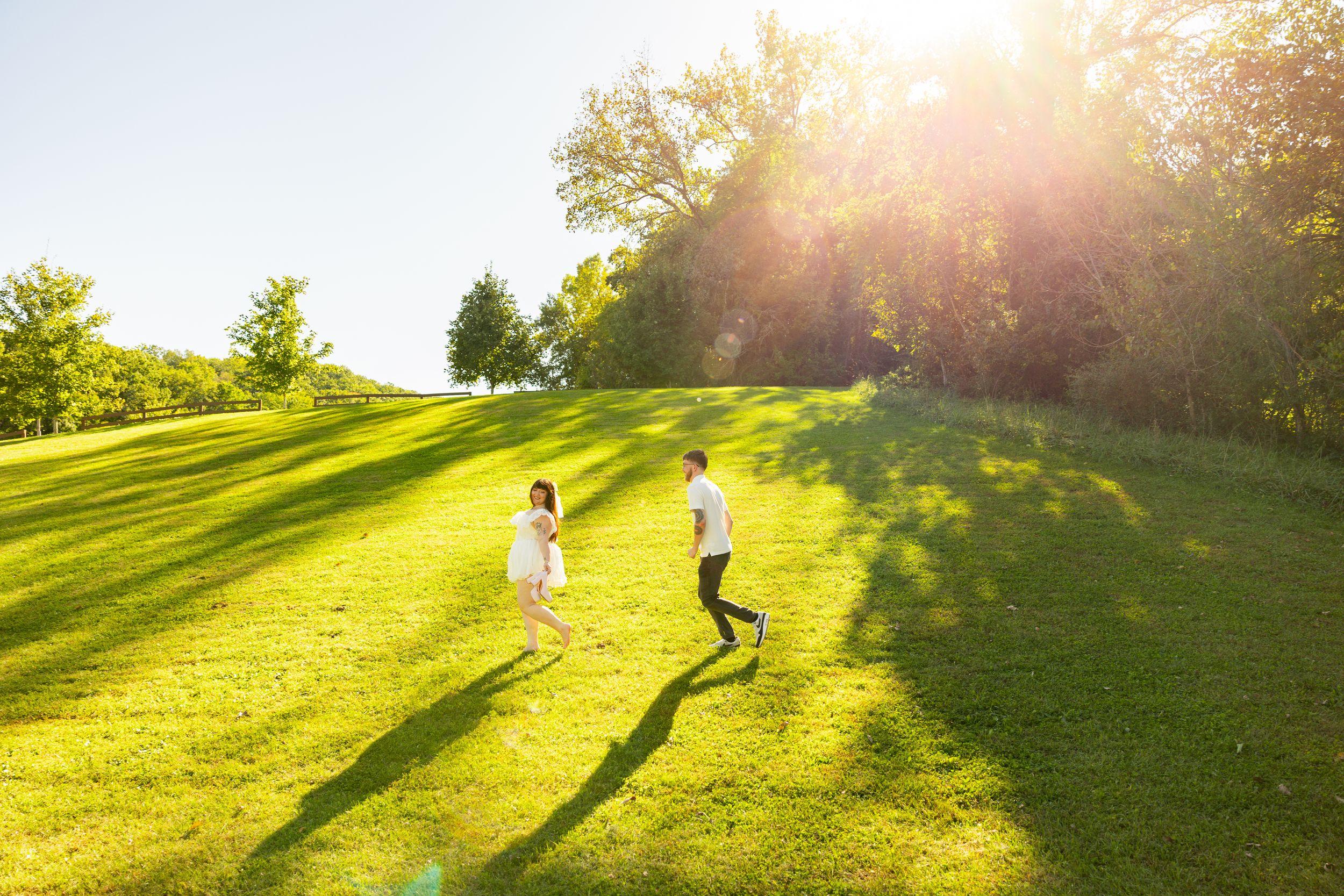 Couple walking hand in hand across a sunlit green grassy hill at golden hour, with long shadows and glowing sun flare through trees.