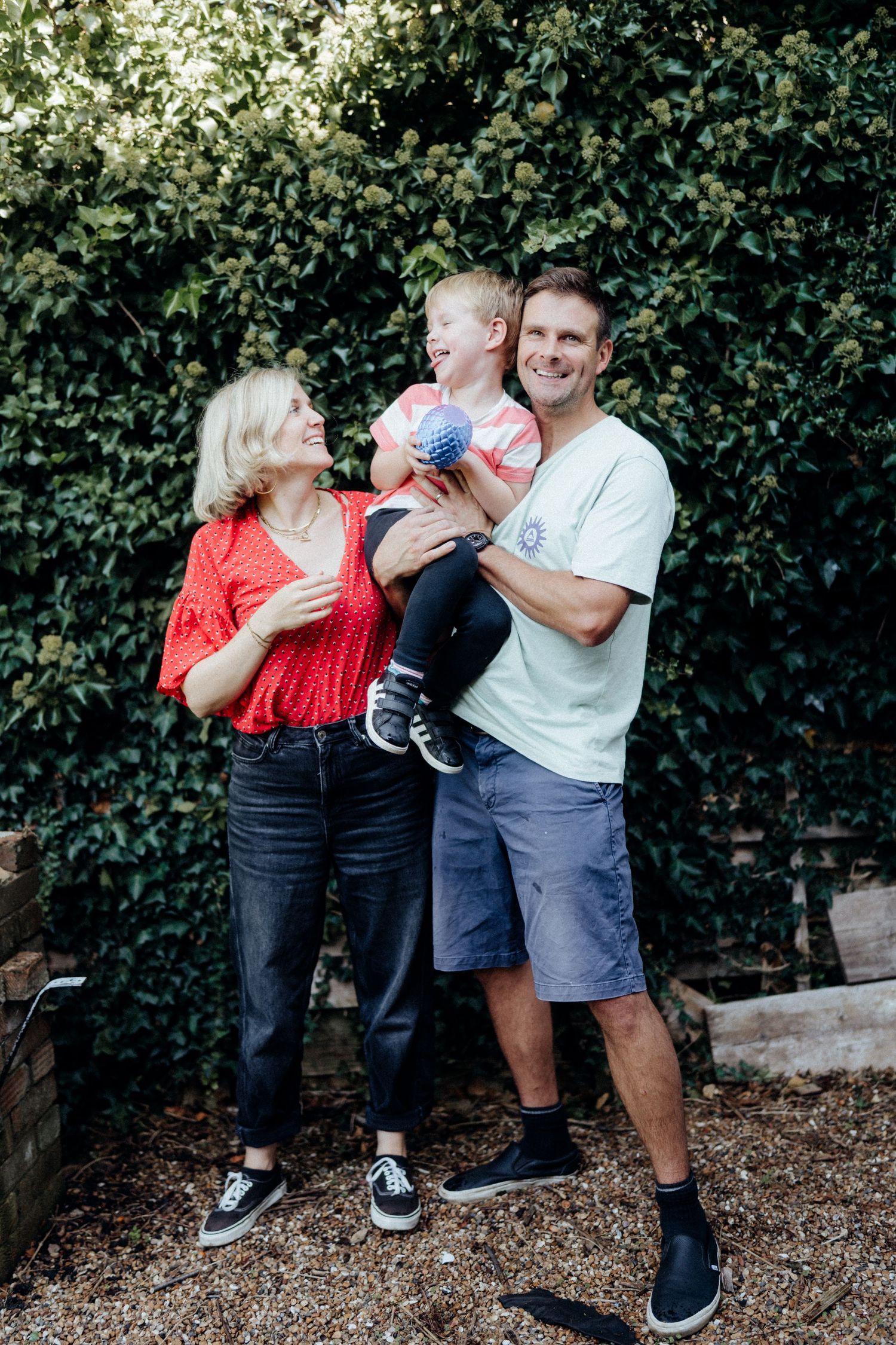 Family shares playful moments together while posing against green hedge background in casual outdoor setting.
