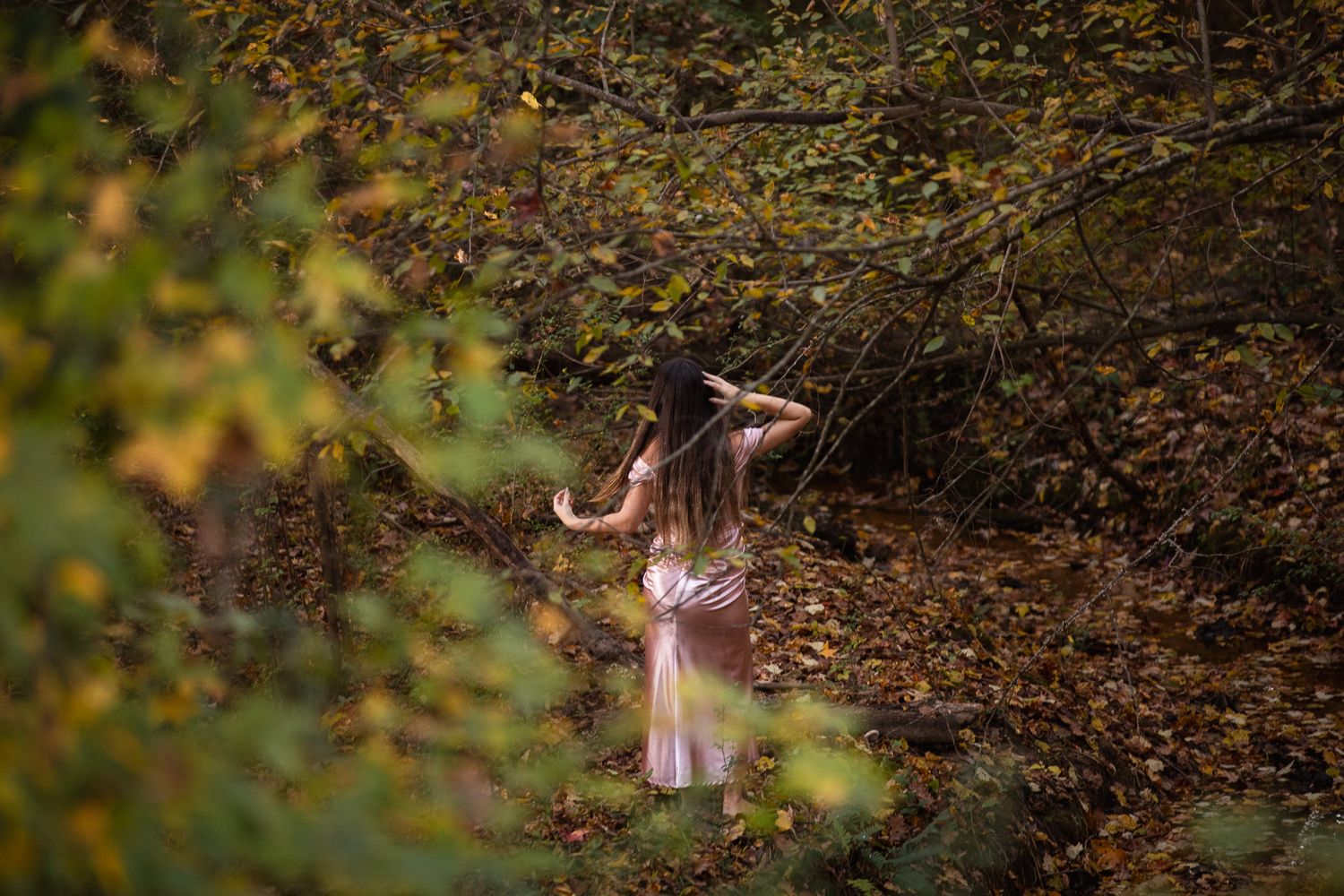 A person in a floral dress poses gracefully in a wooded setting during golden hour lighting.