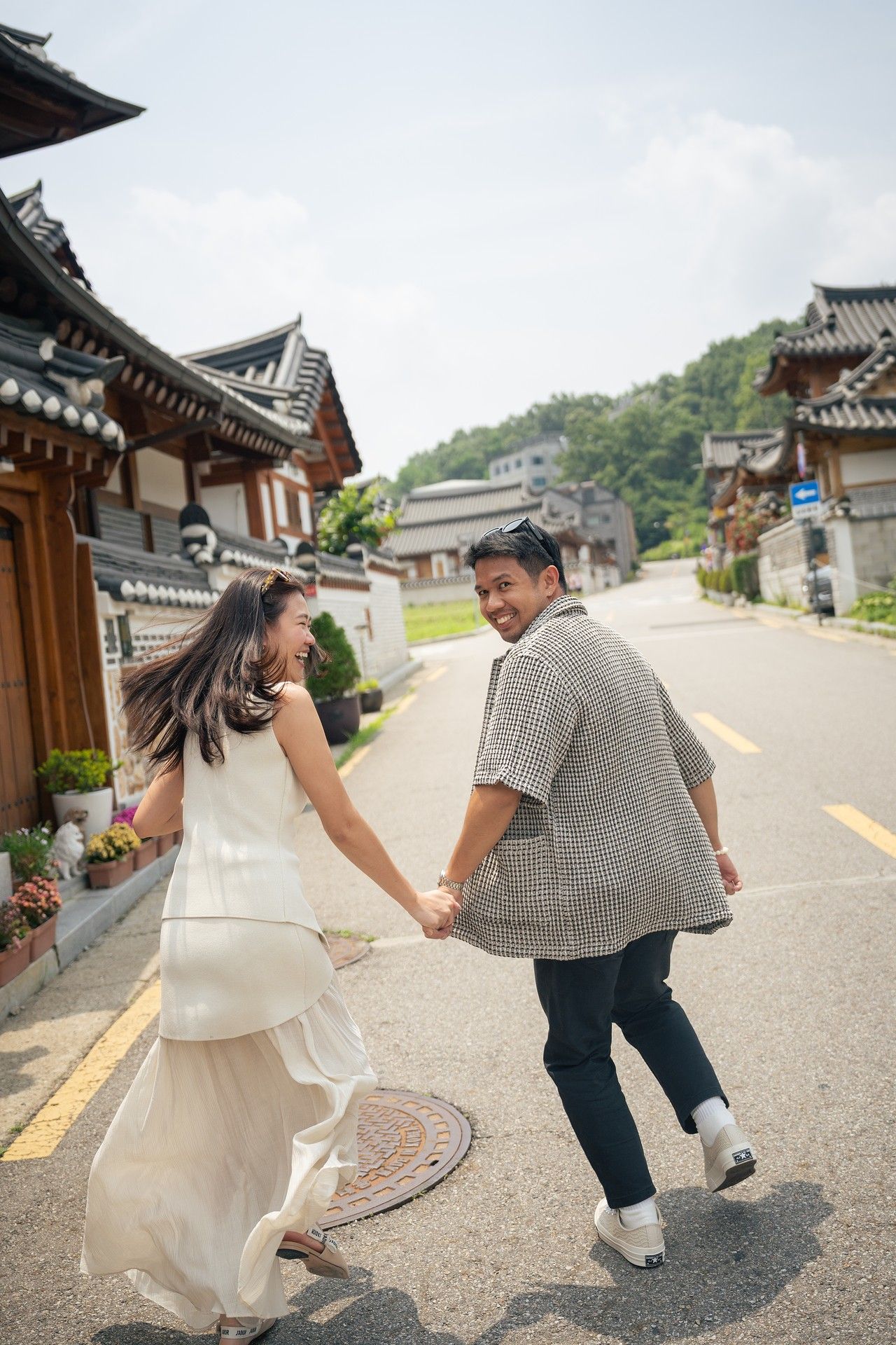 A couple playfully poses for photos on a traditional Korean street lined with historic hanok buildings and architecture.