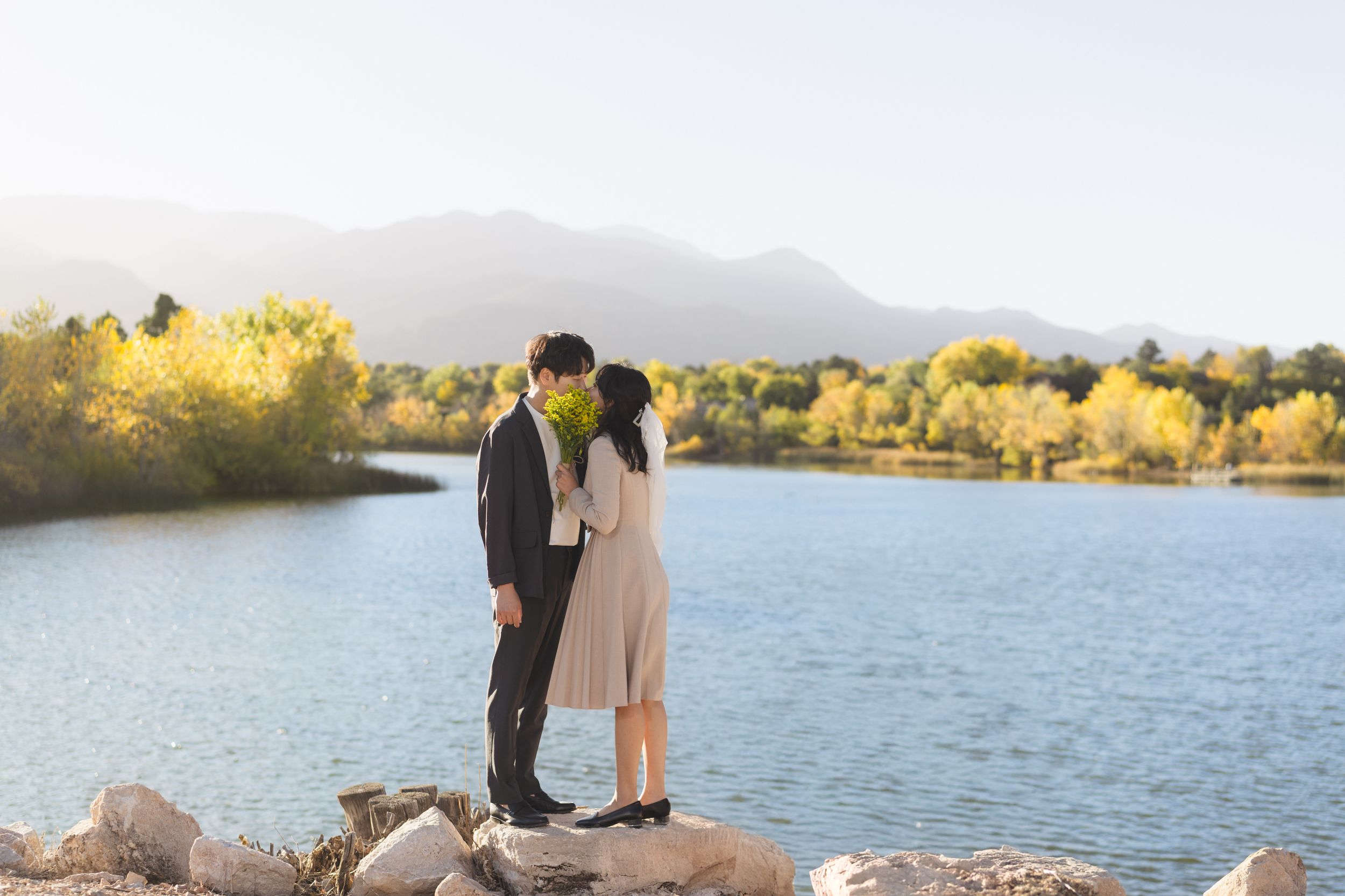 A couple stands together at a scenic lakeside location during golden hour with mountains in the background.