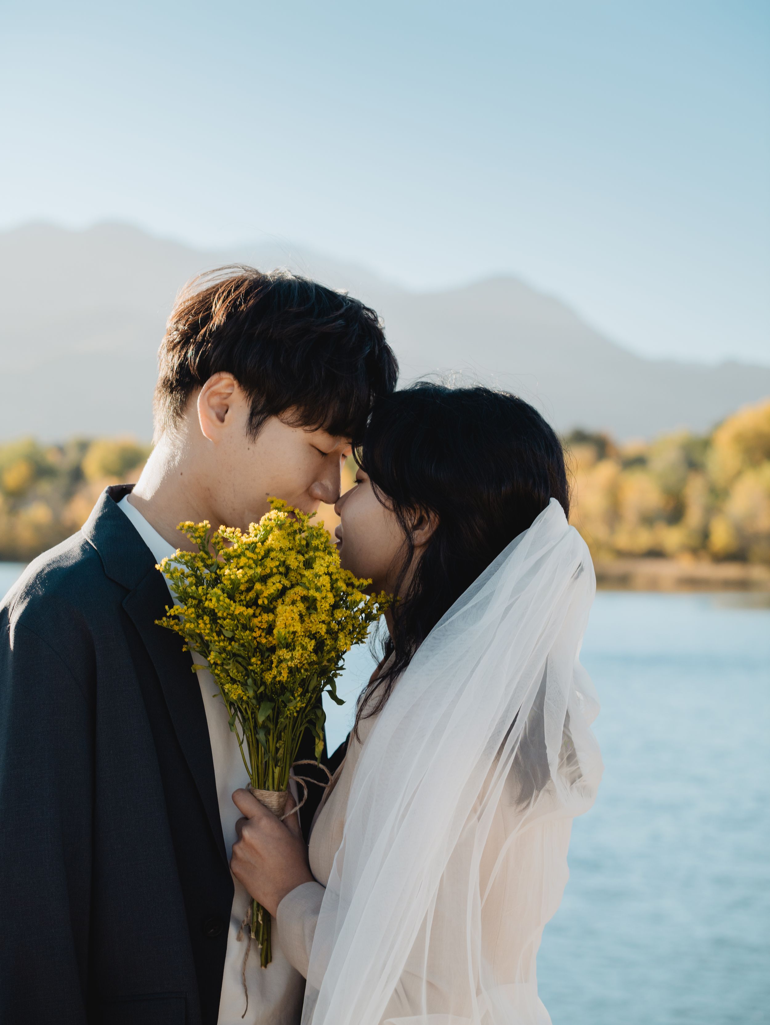 A romantic wedding couple shares an intimate moment by a lake with mountains and fall foliage in the distance.