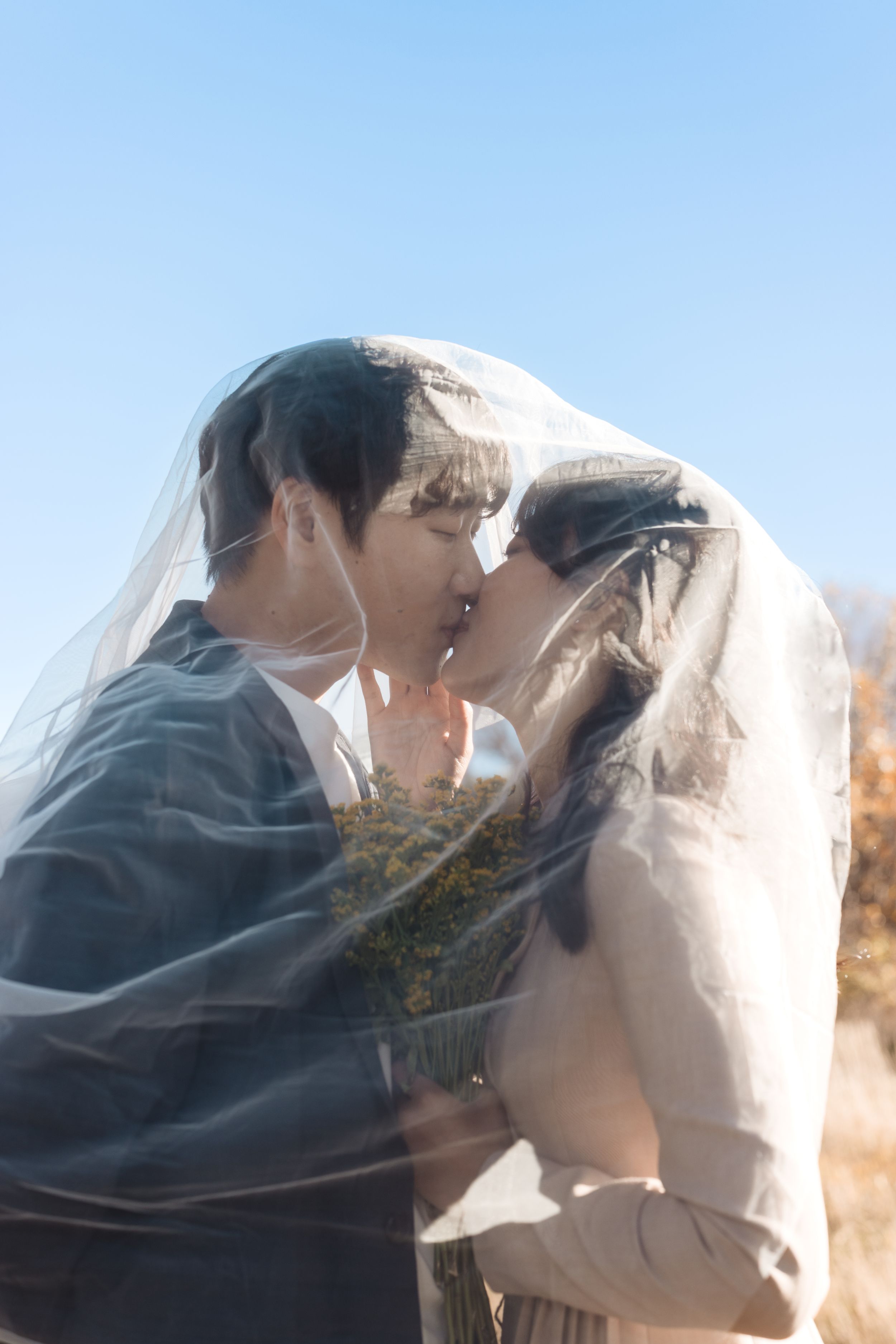 A romantic couple shares intimate kisses under a flowing veil during their outdoor wedding photoshoot.
