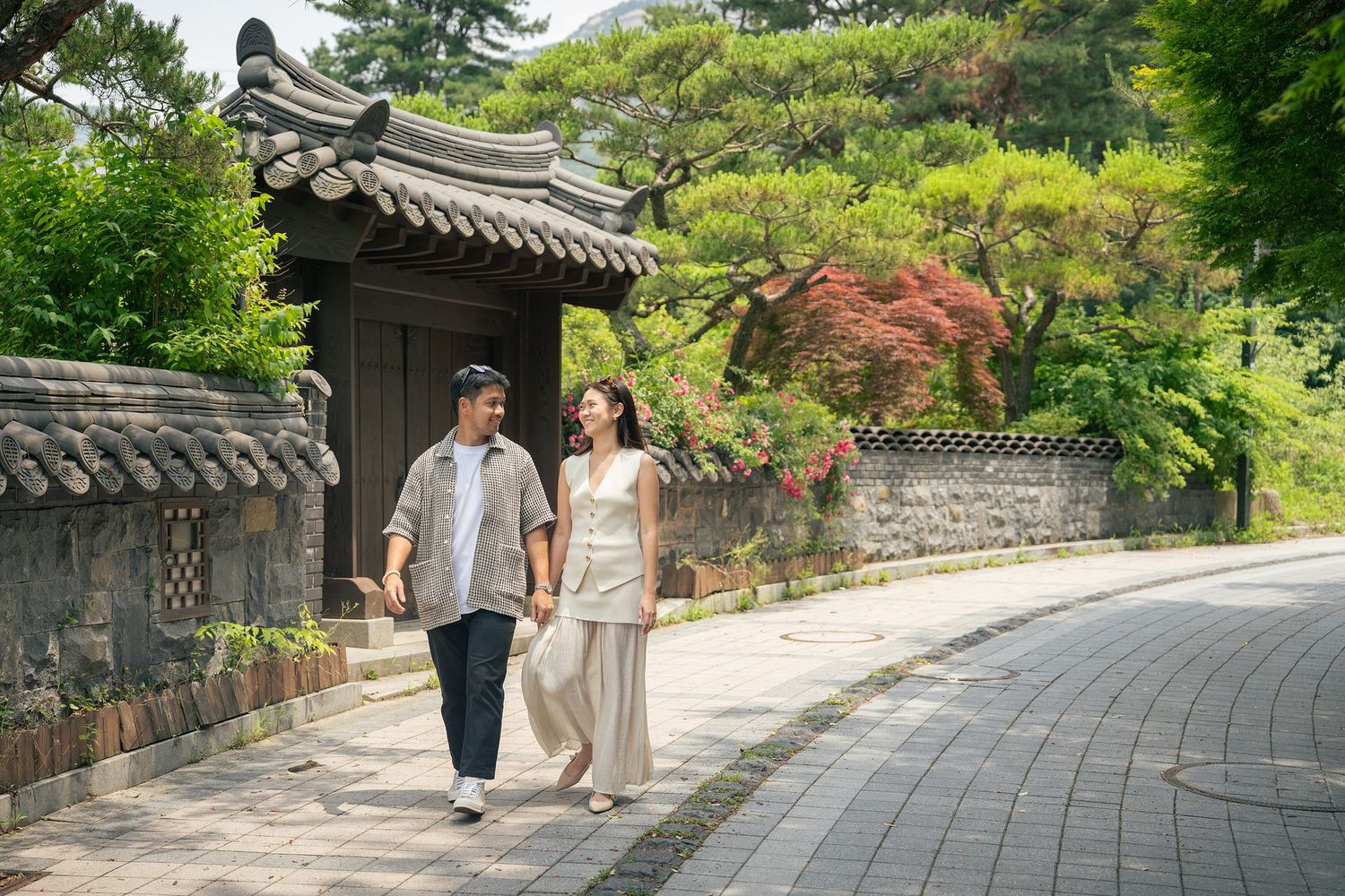 Couple walking along curved stone path by traditional Korean architecture with lush green garden surroundings.