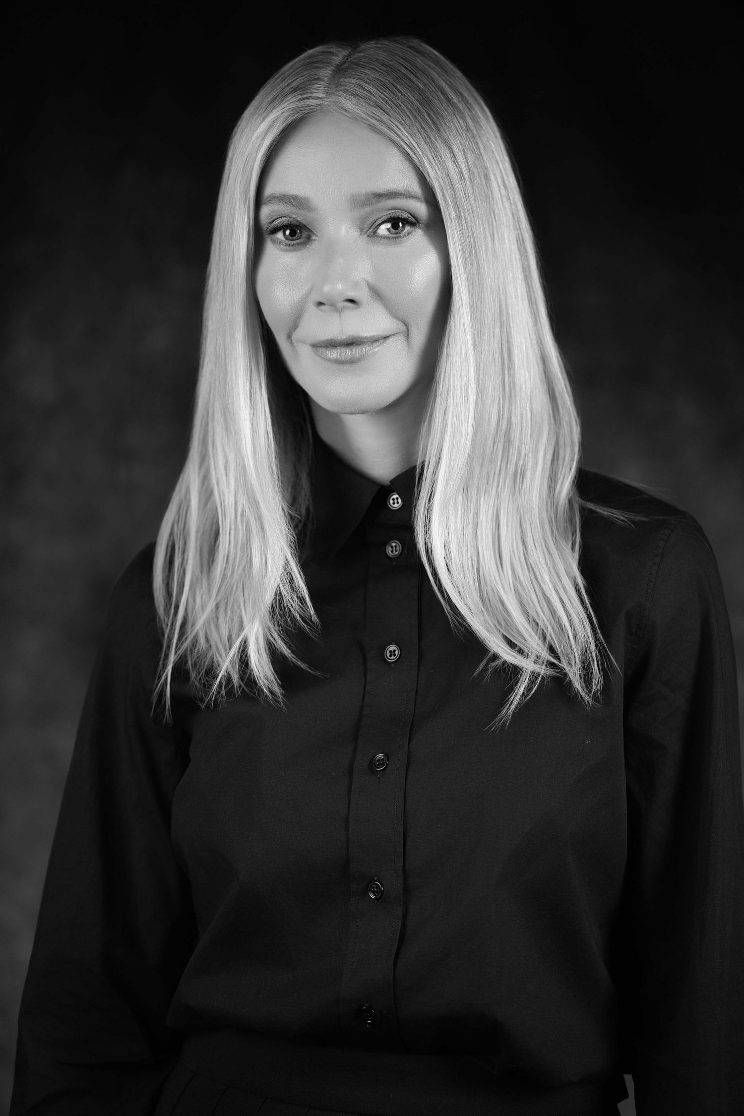 Black and white portrait of a blonde woman in a dark button-up shirt posing against a dark background.