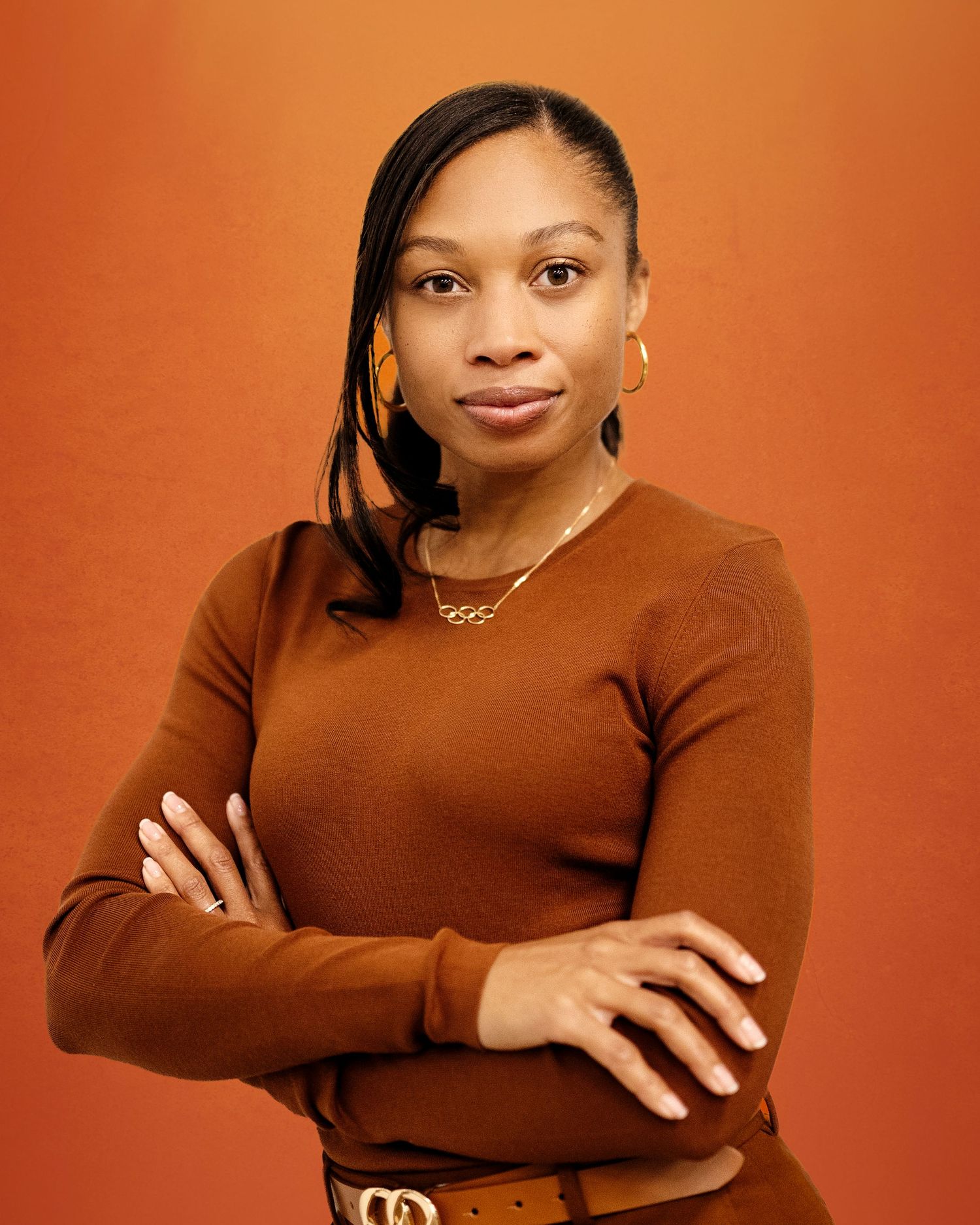 Woman in brown sweater with gold jewelry poses confidently against an orange background.