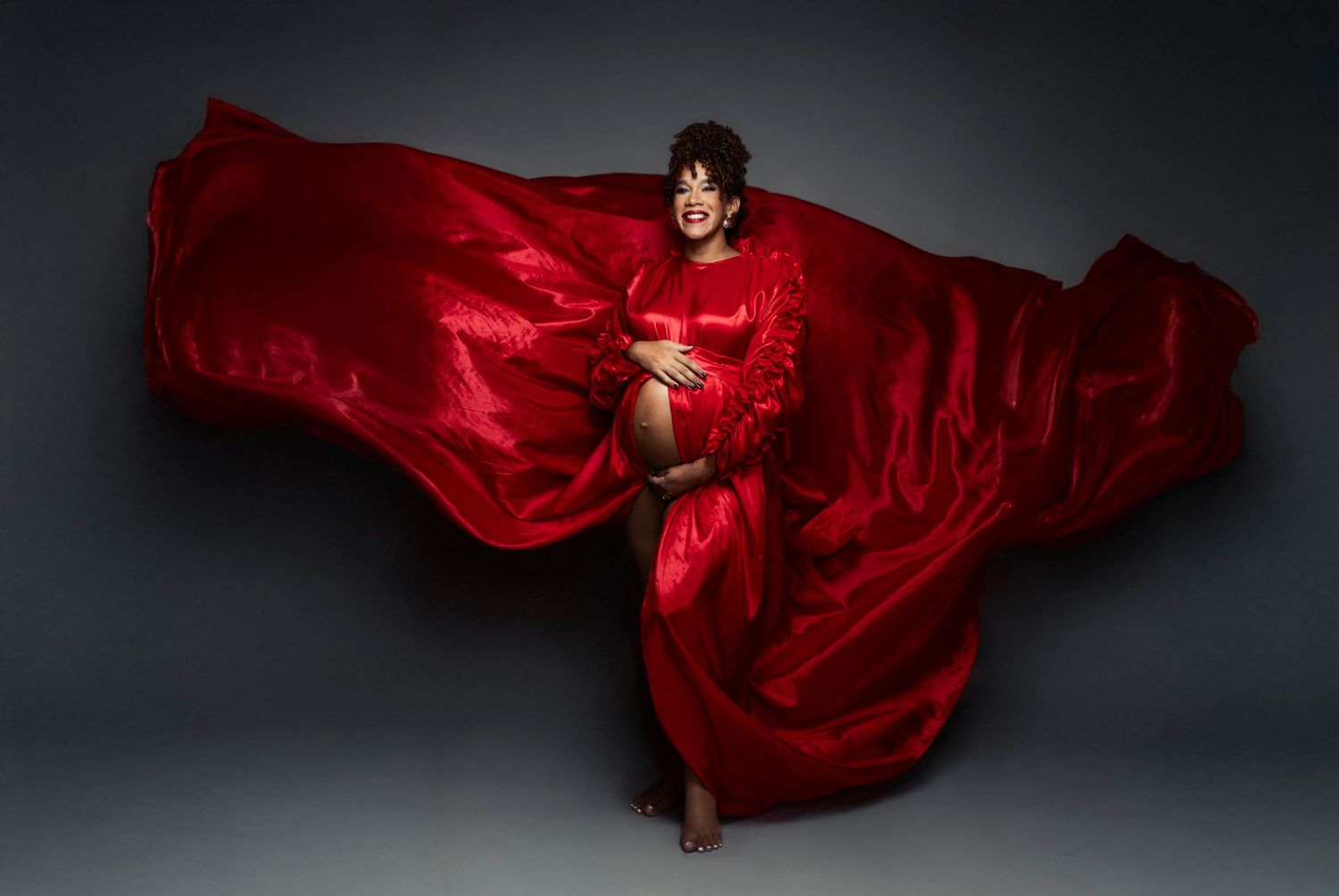 Pregnant woman in flowing red silk gown with dramatic fabric billowing, elegant maternity studio portrait photography.