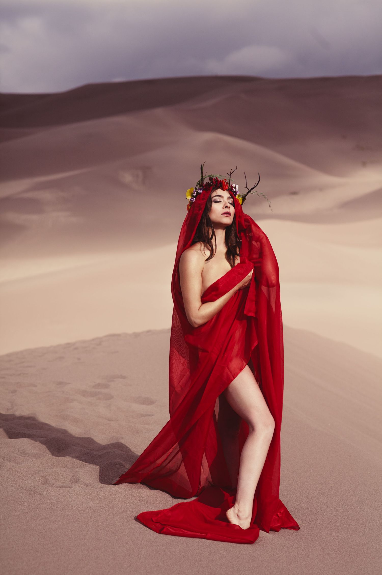 Woman in flowing red fabric and floral antler crown poses gracefully on desert sand dunes under cloudy sky.